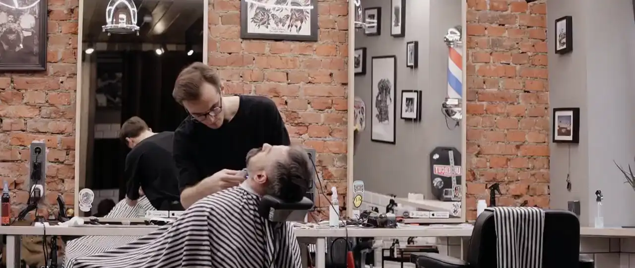 Ein Barbier rasiert einen Gast, der im Stuhl zurückgelehnt liegt; an der Ziegelwand eine Barbierstange