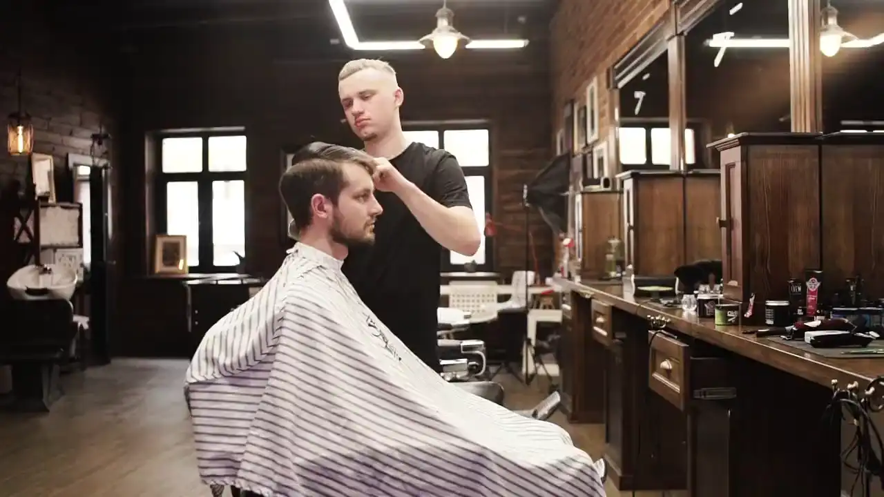 Ein Barbier frisiert einen Gast im gestreiften Umhang, dahinter Spiegel, Holzschränke und hohe Fenster