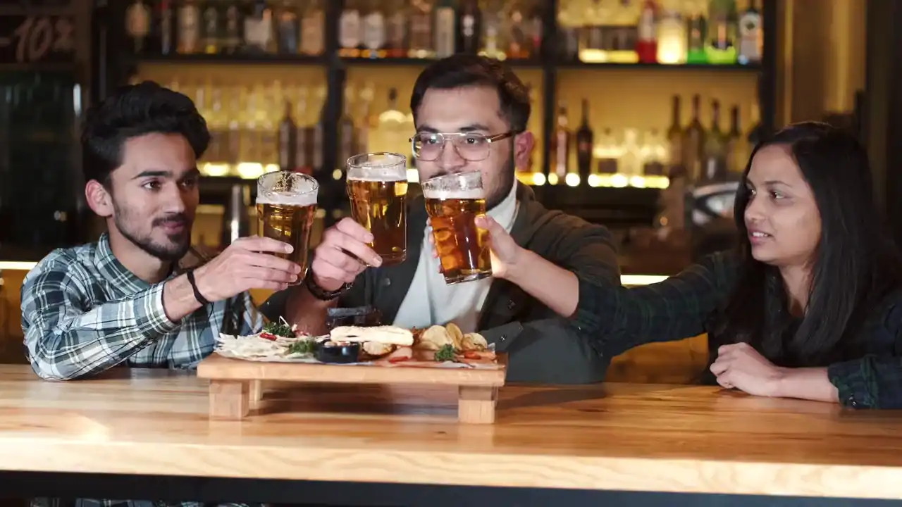 Drei Freunde stoßen an einem Holztisch mit Bier an, vor ihnen ein Brett mit Essen