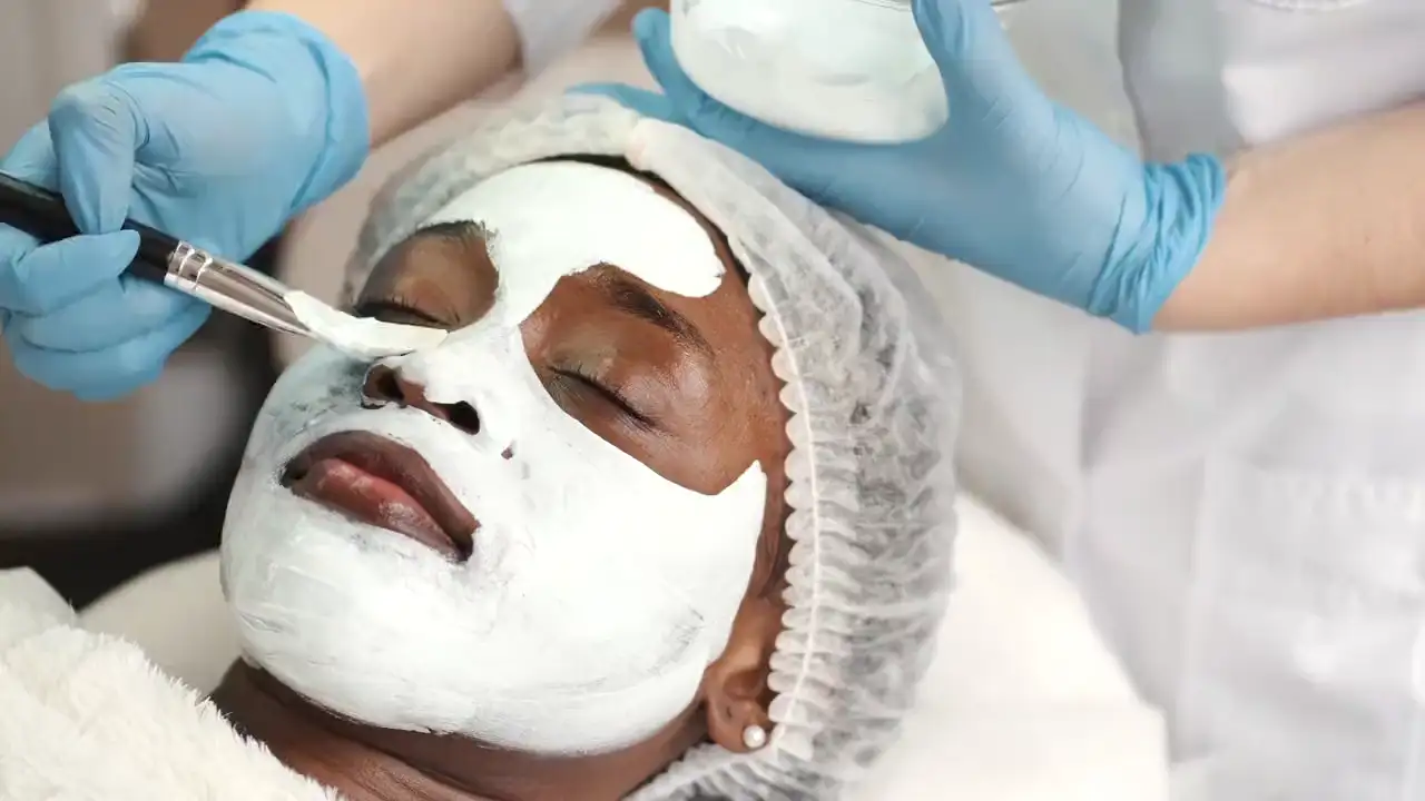 Eine Kosmetikerin in blauen Handschuhen trägt mit einem Pinsel eine weiße Maske auf das Gesicht einer liegenden Kundin auf