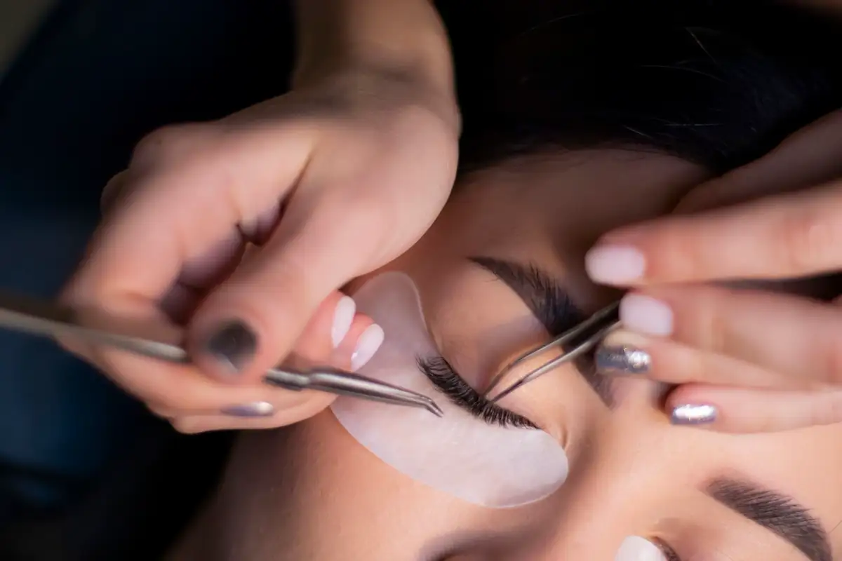 Eine Stylistin setzt mit zwei Pinzetten Wimpern an, unter dem Auge ein Pad