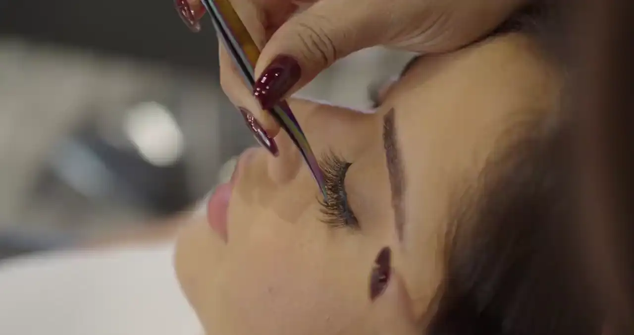 Eine Stylistin setzt mit Pinzette einzelne Wimpern an, die Kundin liegt mit geschlossenen Augen